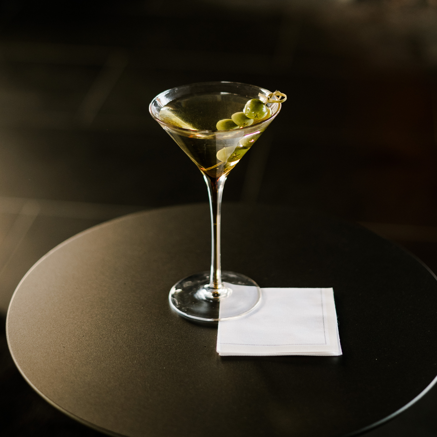 The image shows a martini glass with a clear liquid, likely a martini, garnished with three olives on a toothpick. It’s on a dark table with a white napkin underneath. The dimly lit background highlights the drink as the main focus.