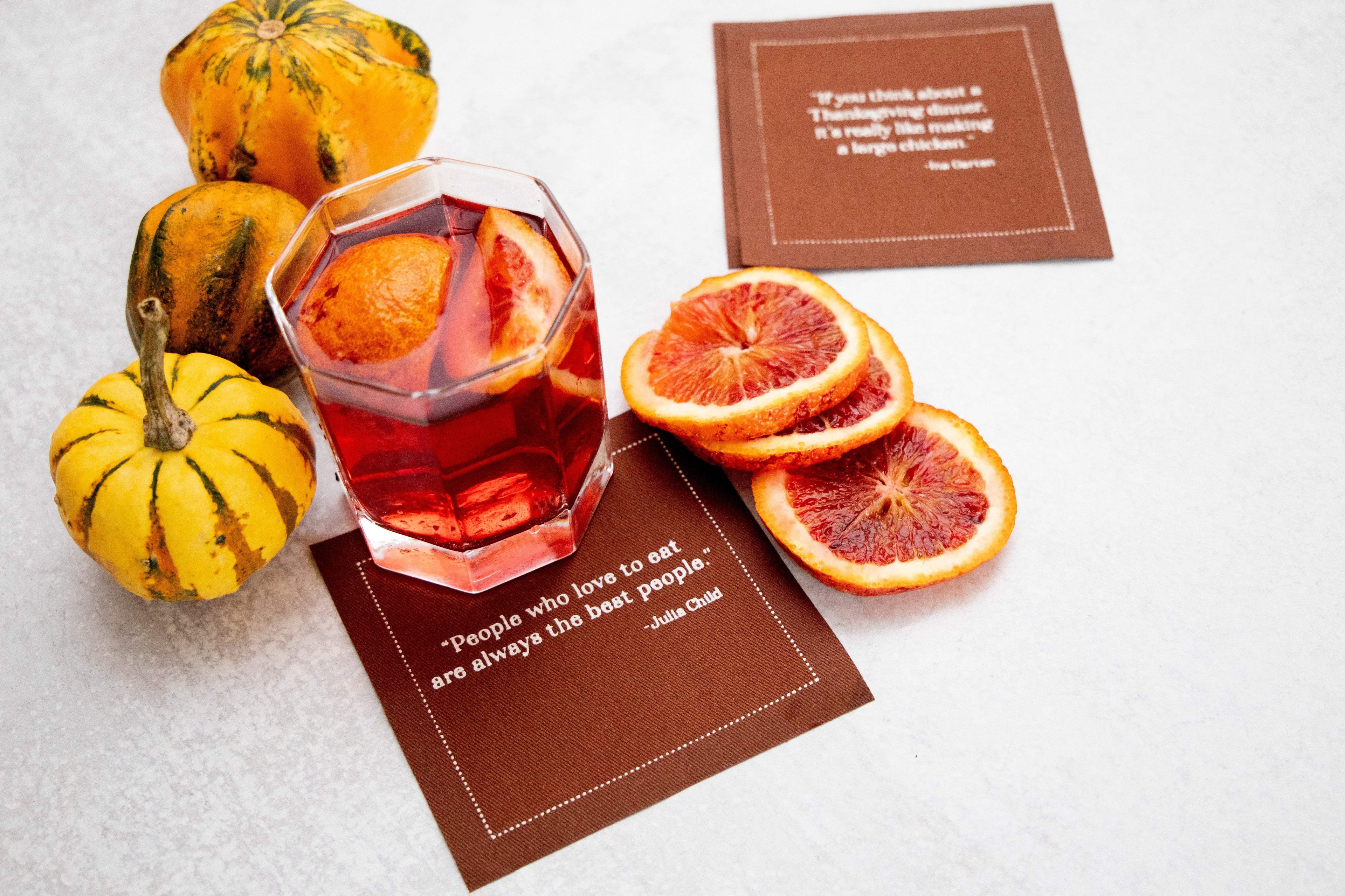 Luxury cocktail napkin with quote, glass of red drink, orange slices, and decorative gourds