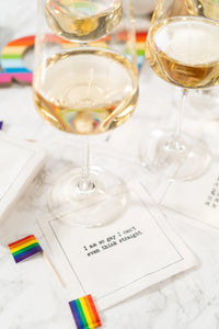 Organic cotton cocktail napkin with printed quote 'I am so gay I can't even think straight' on marble table, rainbow flags and wine glasses visible, party setting.