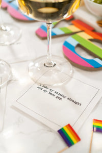 Organic cotton cocktail napkin with printed quote 'I like my whiskey straight and my bars gay' under wine glass, rainbow decor nearby, on a party table setting.