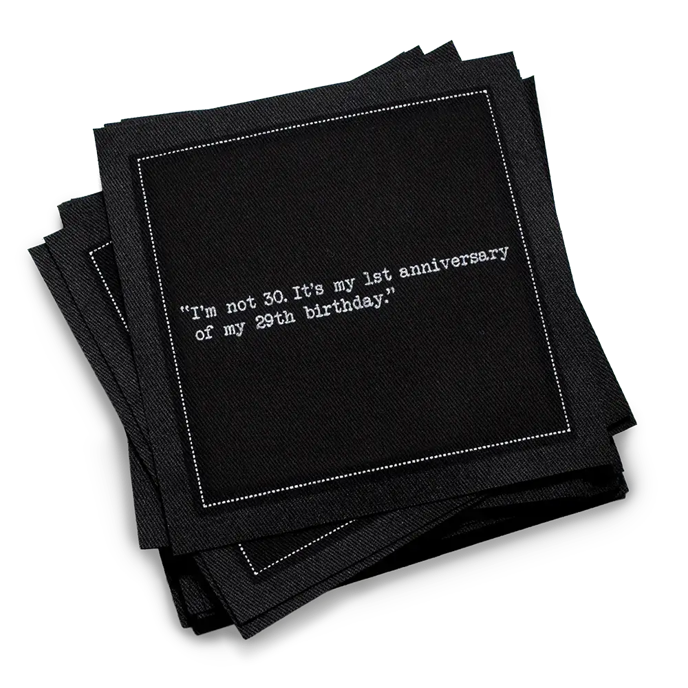 Stack of black 30th birthday quote cocktail napkins with white typewriter-style text reading, “I’m not 30. It’s my 1st anniversary of my 29th birthday.”
