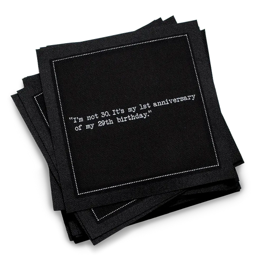 Stack of black 30th birthday quote cocktail napkins with white typewriter-style text reading, “I’m not 30. It’s my 1st anniversary of my 29th birthday.”