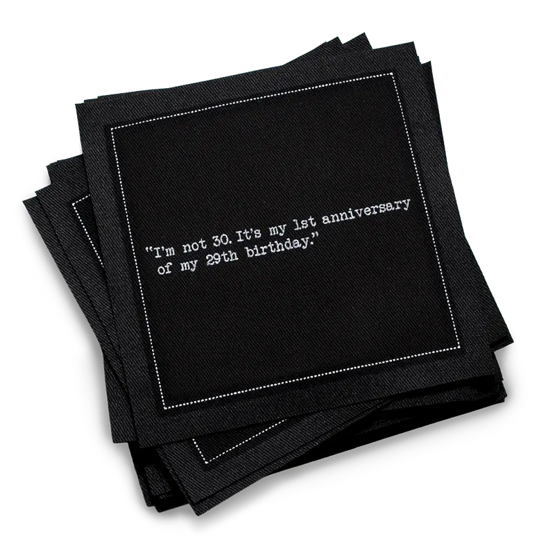 Stack of black 30th birthday quote cocktail napkins with white typewriter-style text reading, “I’m not 30. It’s my 1st anniversary of my 29th birthday.”