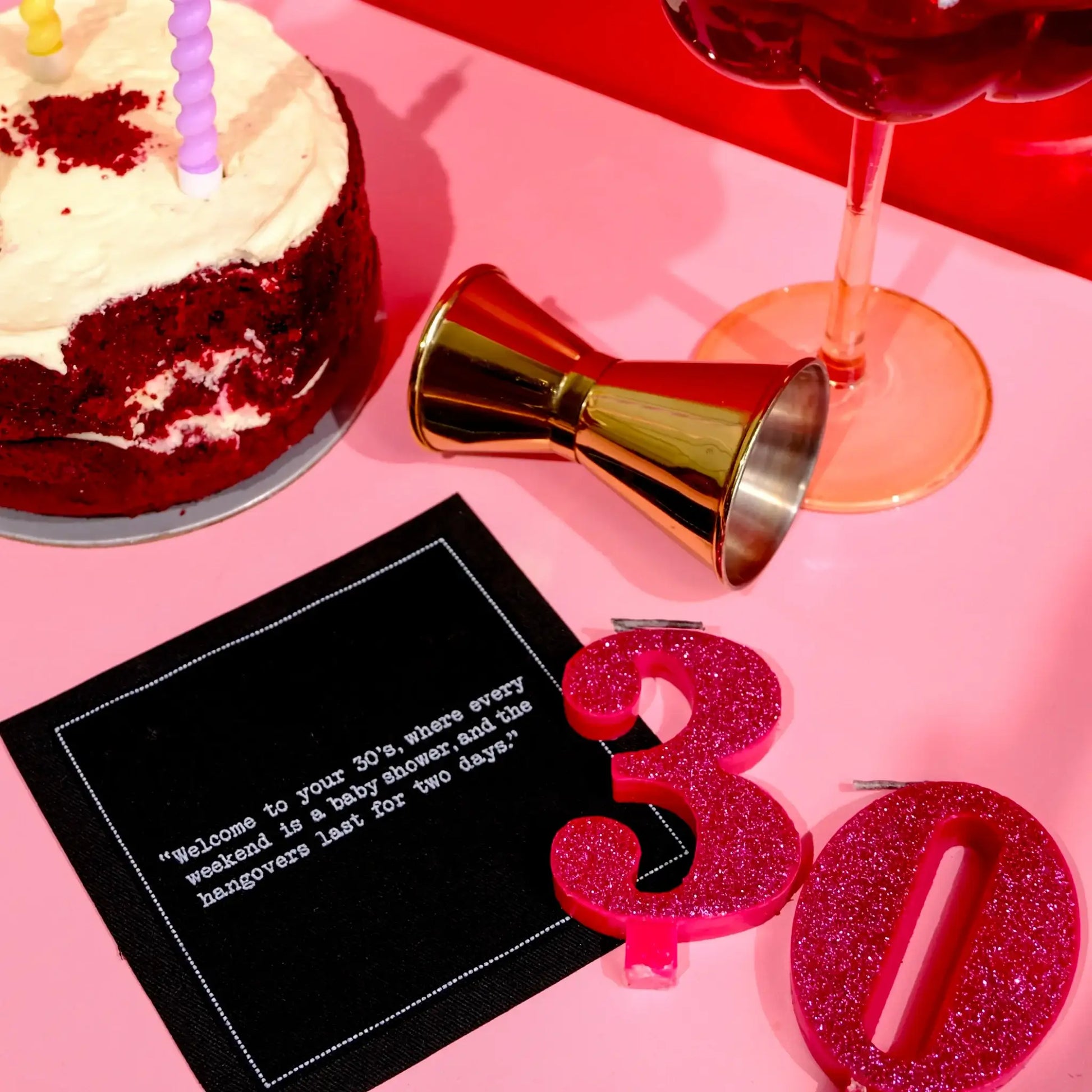 Black 30th birthday quote cocktail napkin with white typewriter-style text reading, “Welcome to your 30s, where every weekend is a baby shower and the hangovers last for two days,” styled with a birthday cake, cocktail, jigger, and glittery “30” candles.