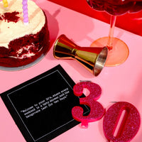 Black 30th birthday quote cocktail napkin with white typewriter-style text reading, “Welcome to your 30s, where every weekend is a baby shower and the hangovers last for two days,” styled with a birthday cake, cocktail, jigger, and glittery “30” candles.