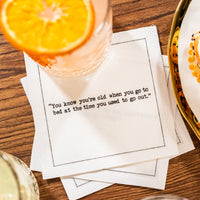 White 30th birthday quote cocktail napkin with black typewriter-style text reading, “You know you’re old when you go to bed at the time you used to go out,” styled beneath a cocktail with an orange slice.