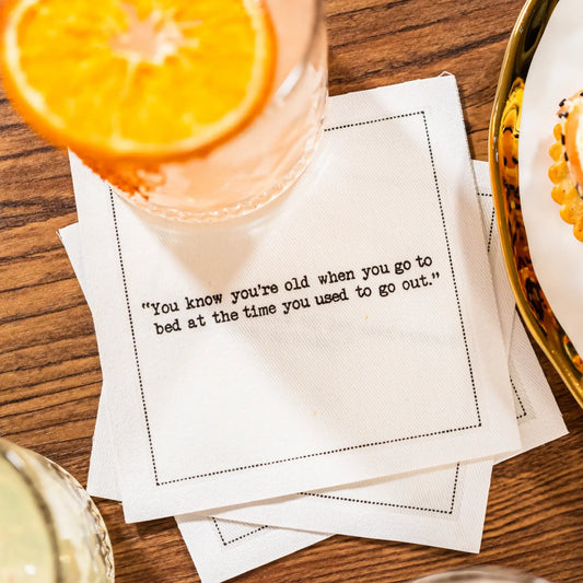 White 30th birthday quote cocktail napkin with black typewriter-style text reading, “You know you’re old when you go to bed at the time you used to go out,” styled beneath a cocktail with an orange slice.