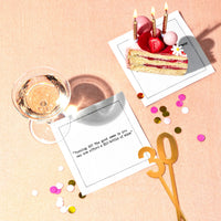 White 30th birthday quote cocktail napkins styled with a birthday cake, wine glass, confetti, and gold “30” decoration, featuring the saying, “Turning 30? The good news is you can now afford a $10 bottle of wine.”