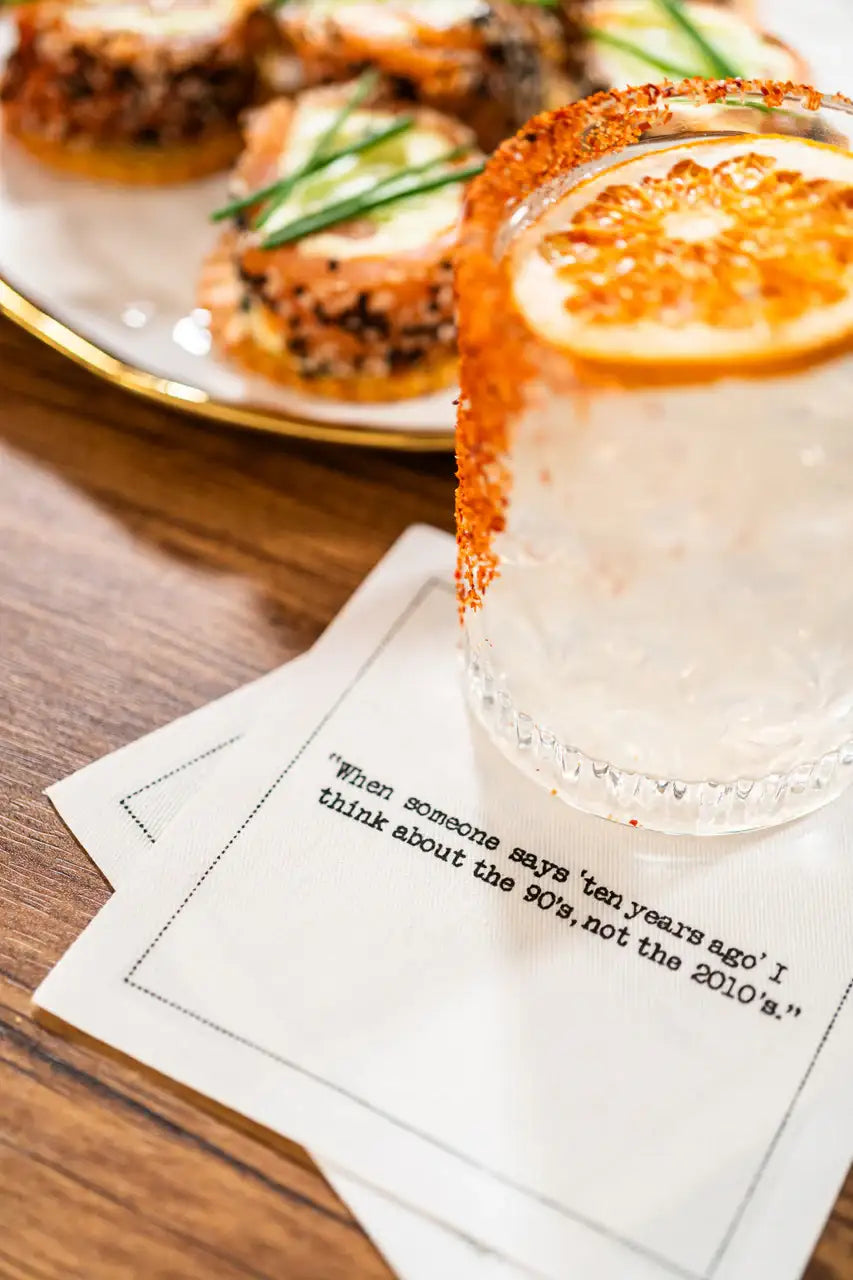 White 30th birthday quote cocktail napkin with black typewriter-style text reading, “When someone says ten years ago I think about the 90’s, not the 2010’s,” styled beneath a citrus cocktail with appetizers nearby.