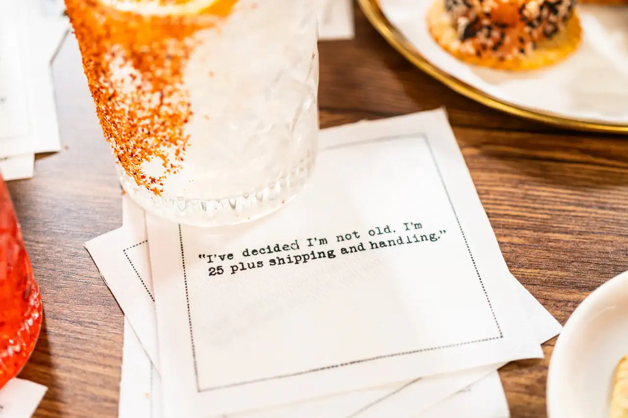 White 30th birthday quote cocktail napkin with black typewriter-style text reading, “I’ve decided I’m not old. I’m 25 plus shipping and handling,” styled beneath a cocktail with appetizers nearby.