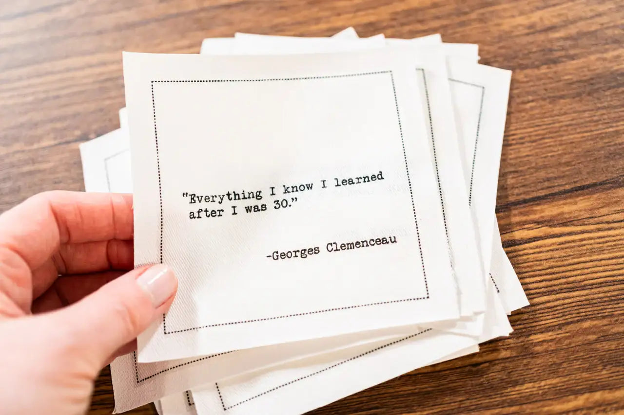 White 30th birthday quote cocktail napkin with black typewriter-style text reading, “Everything I know I learned after I was 30.” – Georges Clemenceau, shown in a stack on a wooden table.