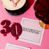 White 30th birthday quote cocktail napkin with black typewriter-style text reading, “They say life begins at 30, but in your case it’s more like a new chapter in your book of secrets,” styled with a birthday cake, drink, and glittery “30” candles.