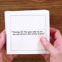 Hands holding a stack of white 30th birthday quote cocktail napkins with black typewriter-style text reading, “Turning 30? The good news is you can now afford a $10 bottle of wine.”