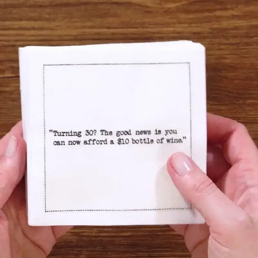Hands holding a stack of white 30th birthday quote cocktail napkins with black typewriter-style text reading, “Turning 30? The good news is you can now afford a $10 bottle of wine.”