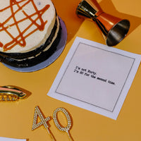 Organic cotton cocktail napkin printed with the quote "I'm not Forty. I'm 20 for the second time," styled on an orange table with a birthday cake, gold bar tools, and a sparkly "40" cake topper, from Five Star Napkins' 40th Birthday Quotes collection