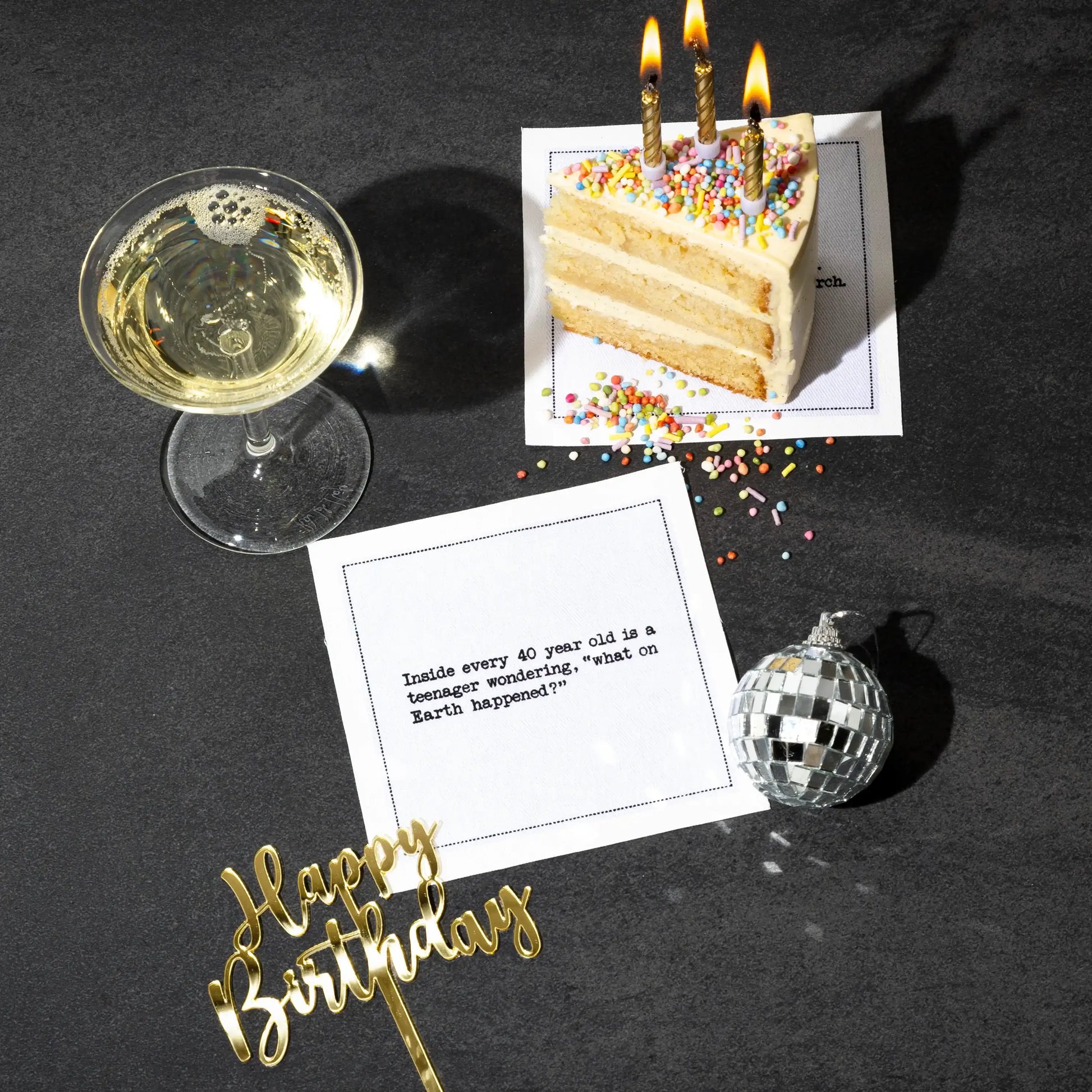 Organic cotton cocktail napkin printed with the quote "Inside every 40 year old is a teenager wondering, 'what on Earth happened?'" styled on a dark table with birthday cake, champagne, a disco ball, and a "Happy Birthday" cake topper, from Five Star Napkins' 40th Birthday Quotes collection