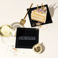 Organic cotton cocktail napkin, black, printed with the quote "40 is the perfect age. You're old enough to recognize your mistakes but young enough to make some more," styled with birthday cake, champagne, and a disco ball ornament, from Five Star Napkins' 40th Birthday Quotes collection