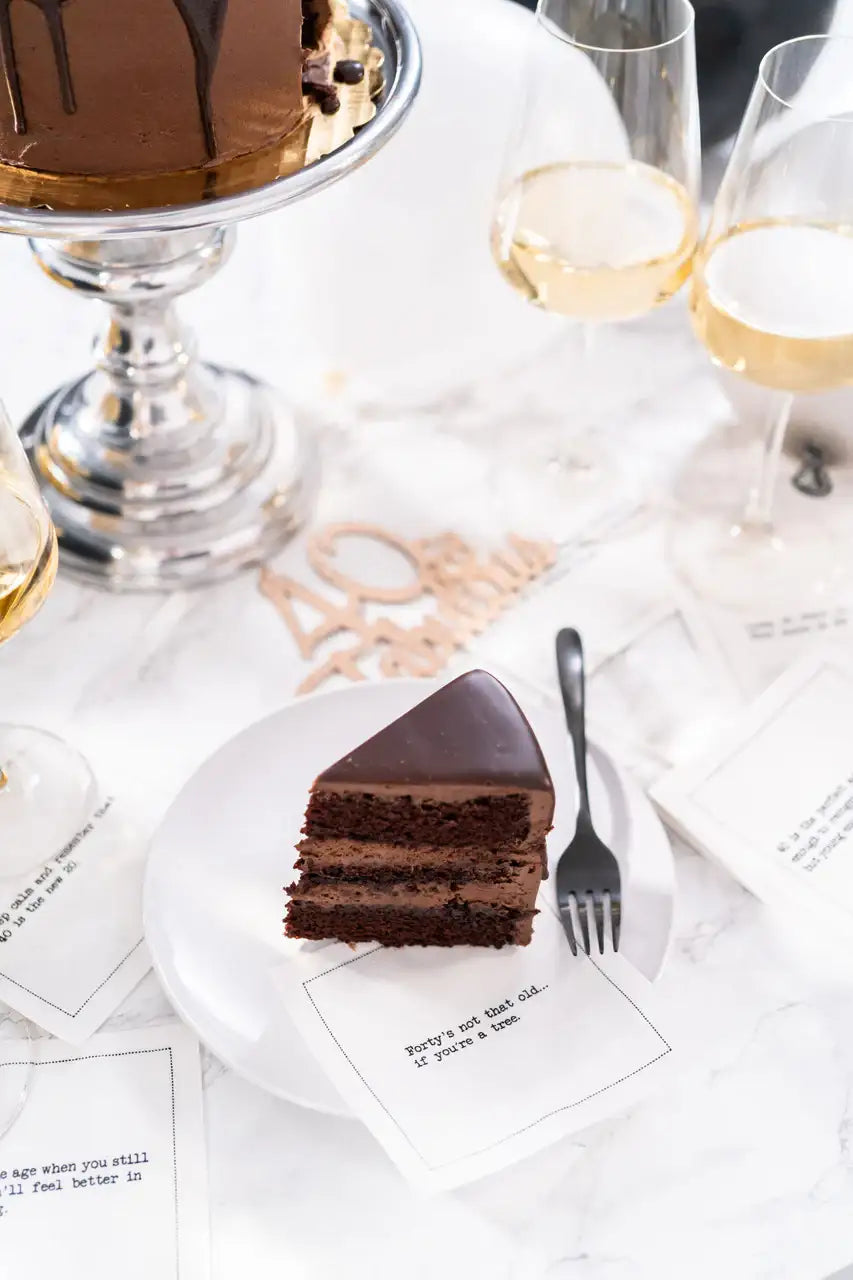 Organic cotton cocktail napkin printed with the quote "Forty's not that old... if you're a tree," styled on a marble table with a chocolate cake, champagne glasses, and rose gold "40" decor, from Five Star Napkins' 40th Birthday Quotes collection