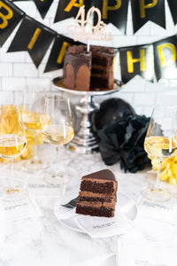 Organic cotton cocktail napkins printed with 40th birthday quotes, styled on a marble table with a chocolate birthday cake, wine glasses, and a "Happy 40th Birthday" banner, from Five Star Napkins' 40th Birthday Quotes collection