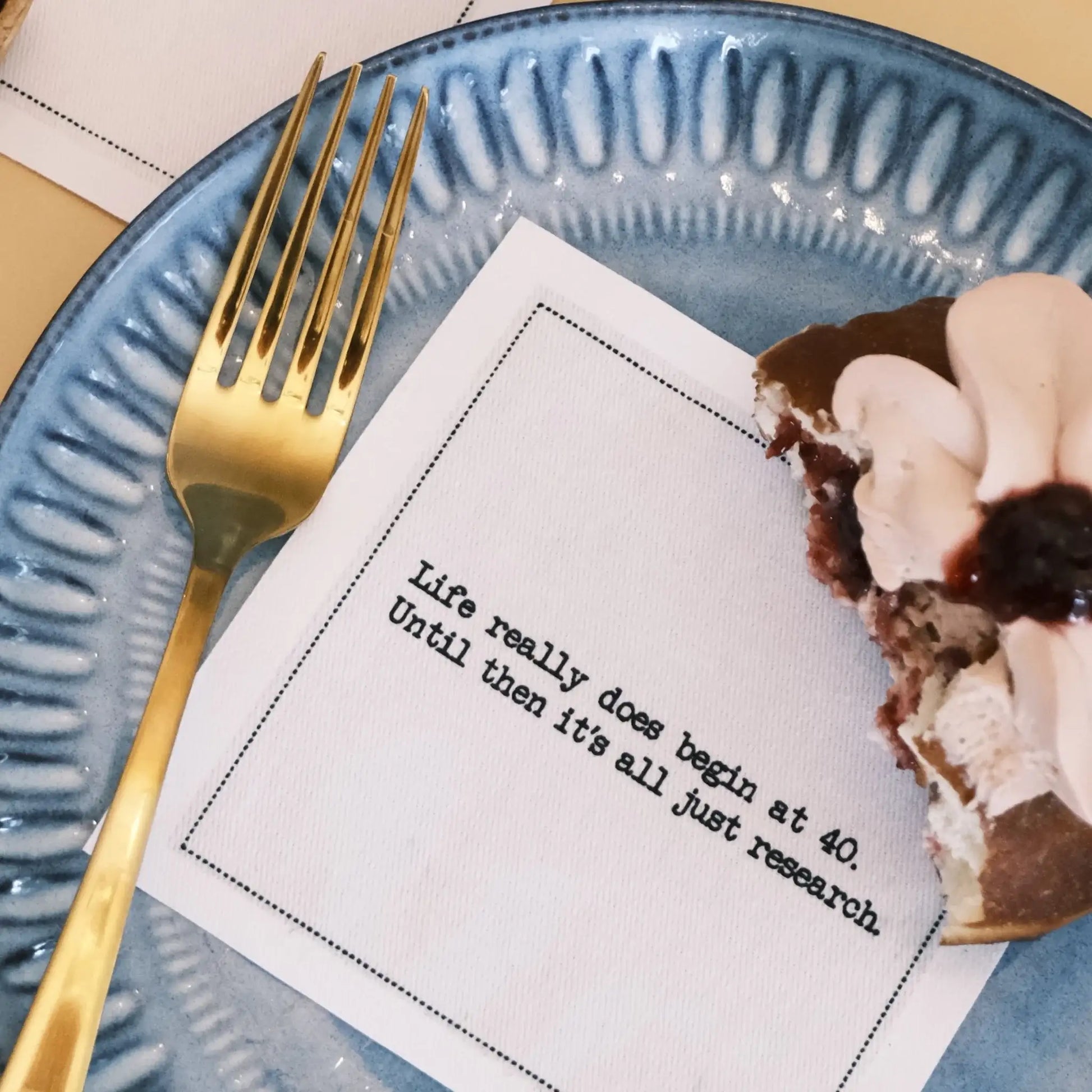 Organic cotton cocktail napkin printed with the quote "Life really does begin at 40. Until then it's all just research," styled on a blue plate with a slice of dessert and a gold fork, from Five Star Napkins' 40th Birthday Quotes collection
