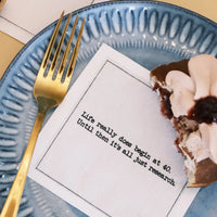 Organic cotton cocktail napkin printed with the quote "Life really does begin at 40. Until then it's all just research," styled on a blue plate with a slice of dessert and a gold fork, from Five Star Napkins' 40th Birthday Quotes collection