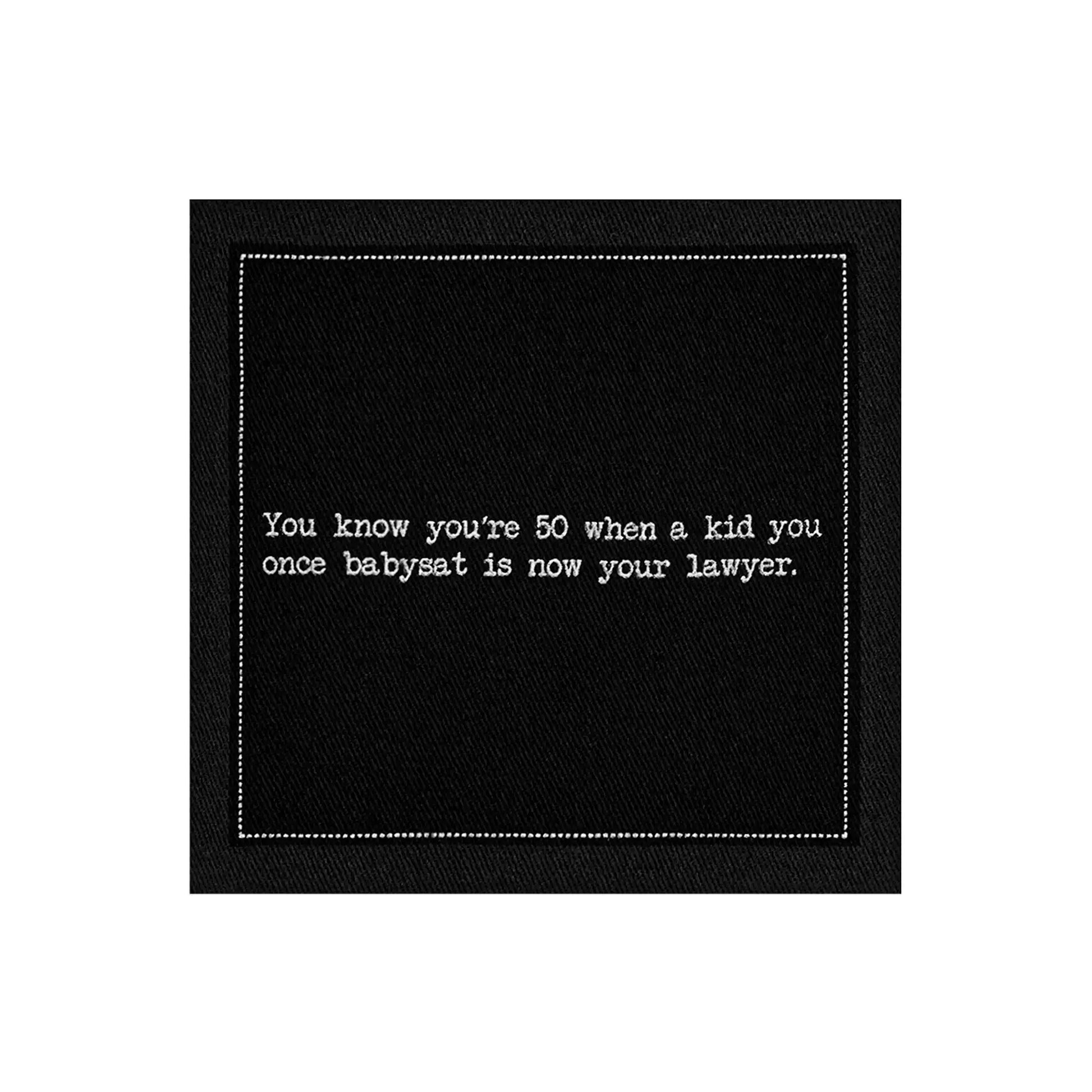 Organic cotton cocktail napkin, black, with white dotted border printed with the quote "You know you're 50 when a kid you once babysat is now your lawyer," from Five Star Napkins' 50th Birthday Quotes collection