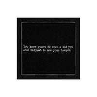 Organic cotton cocktail napkin, black, with white dotted border printed with the quote "You know you're 50 when a kid you once babysat is now your lawyer," from Five Star Napkins' 50th Birthday Quotes collection