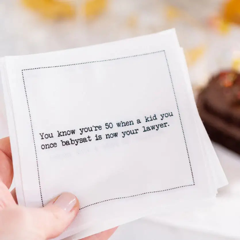 Organic cotton cocktail napkin printed with the quote "You know you're 50 when a kid you once babysat is now your lawyer," held in hand with a chocolate cake in the background, from Five Star Napkins' 50th Birthday Quotes collection