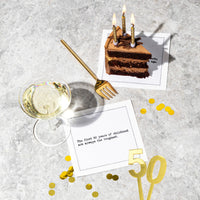 Organic cotton cocktail napkin printed with the quote "The first 50 years of childhood are always the toughest," styled on a marble table with a slice of chocolate cake, a glass of wine, gold confetti, and a gold "50" decoration, from Five Star Napkins' 50th Birthday Quotes collection