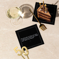 Organic cotton cocktail napkin, black, printed with the quote "The best part of being over fifty, is that you did most of your stupid stuff before the internet," styled on a marble table with a slice of chocolate cake, a glass of champagne, and a gold "50" decoration, from Five Star Napkins' 50th Birthday Quotes collection