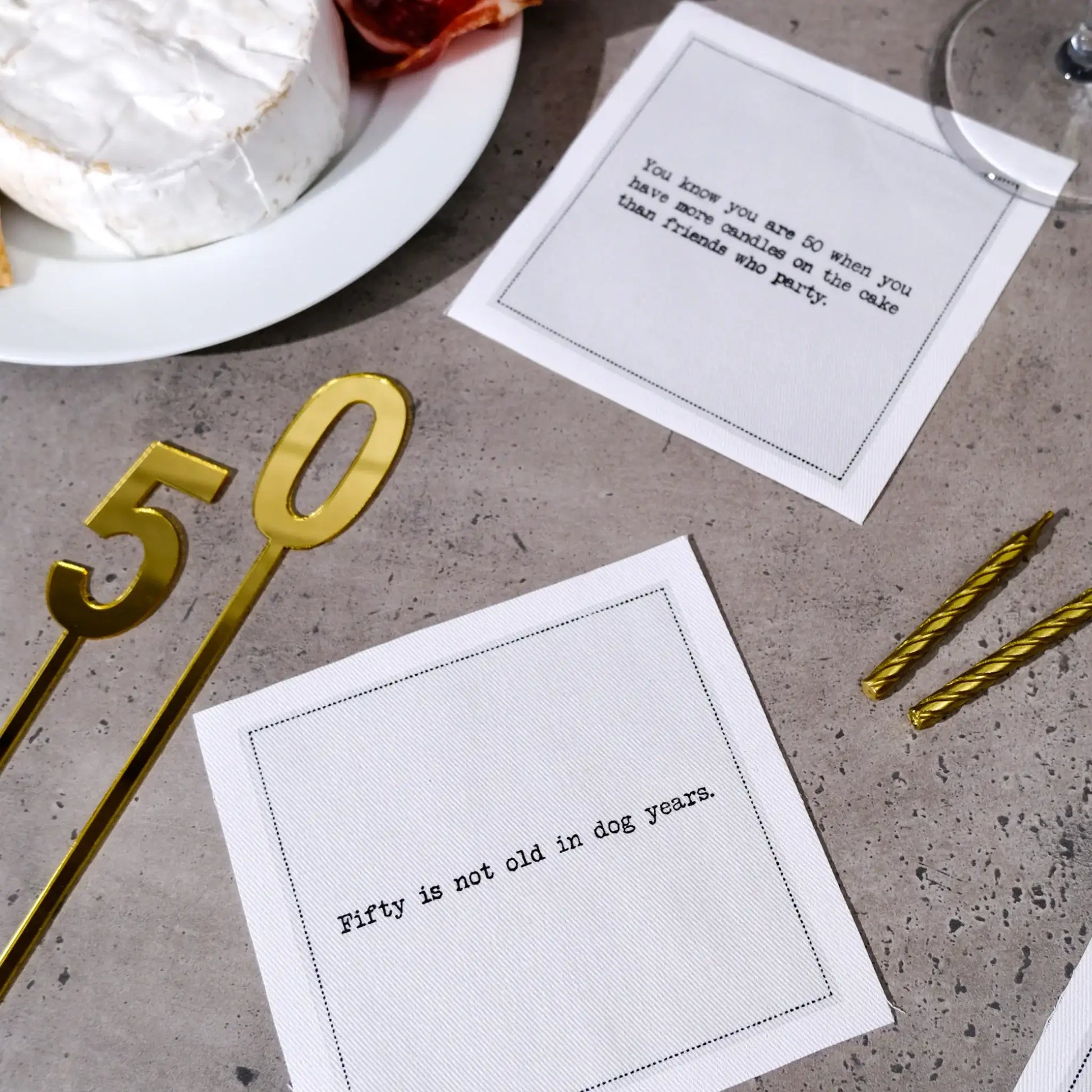 Organic cotton cocktail napkins printed with the quotes "You know you are 50 when you have more candles on the cake than friends who party" and "Fifty is not old in dog years," styled on a stone table with brie cheese, gold candles, and a gold "50" decoration, from Five Star Napkins' 50th Birthday Quotes collection