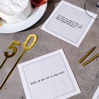 Organic cotton cocktail napkins printed with the quotes "You know you are 50 when you have more candles on the cake than friends who party" and "Fifty is not old in dog years," styled on a stone table with brie cheese, gold candles, and a gold "50" decoration, from Five Star Napkins' 50th Birthday Quotes collection
