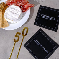 Organic cotton cocktail napkins, black, printed with the quotes "Don't worry about being 50. You are still going to make mistakes, only slower." and "If you haven't grown up by age 50, you don't have to," styled on a stone table with a charcuterie and cheese board and a gold "50" decoration, from Five Star Napkins' 50th Birthday Quotes collection