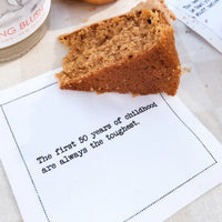 Organic cotton cocktail napkin printed with the quote "The first 50 years of childhood are always the toughest," styled under a slice of cake on a linen tablecloth, from Five Star Napkins' 50th Birthday Quotes collection