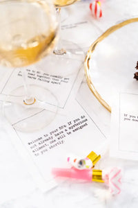 Organic cotton cocktail napkins printed with 50th birthday quotes, including "Welcome to your 50's. If you do not already have a mysterious ailment, one will be assigned to you shortly," styled on a marble table with champagne glasses and party blowers, from Five Star Napkins' 50th Birthday Quotes collection