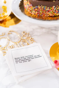 Organic cotton cocktail napkin with 'Happy 50th birthday! You have now reached the age where compliments are followed by for your age.' printed quote, styled at a birthday party table with chocolate sprinkle cake.