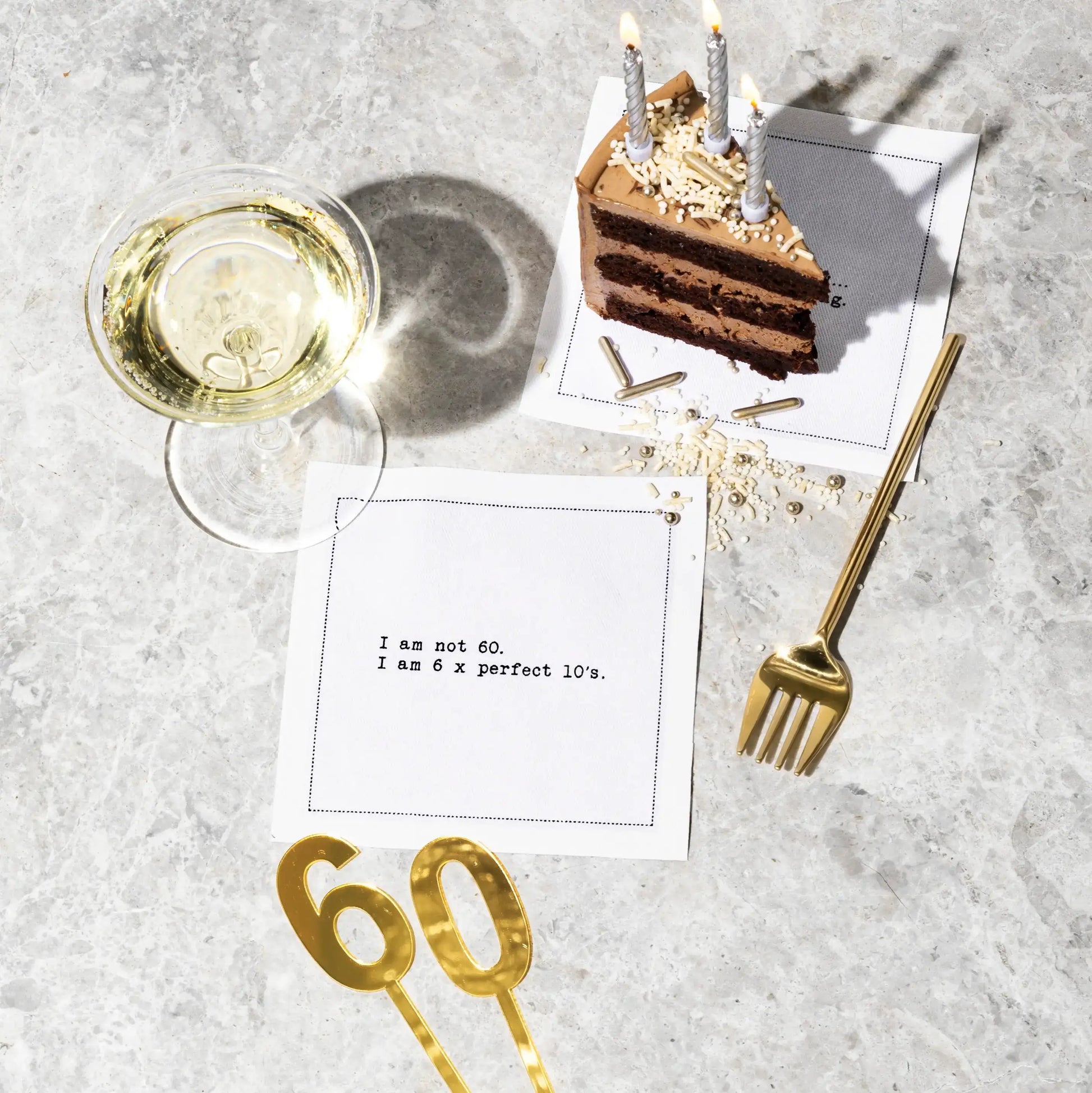 Organic cotton cocktail napkin printed with the quote "I am not 60. I am 6 x perfect 10's," styled on a marble table with chocolate cake, champagne, and gold "60" cake toppers, from Five Star Napkins' 60th birthday quote collection