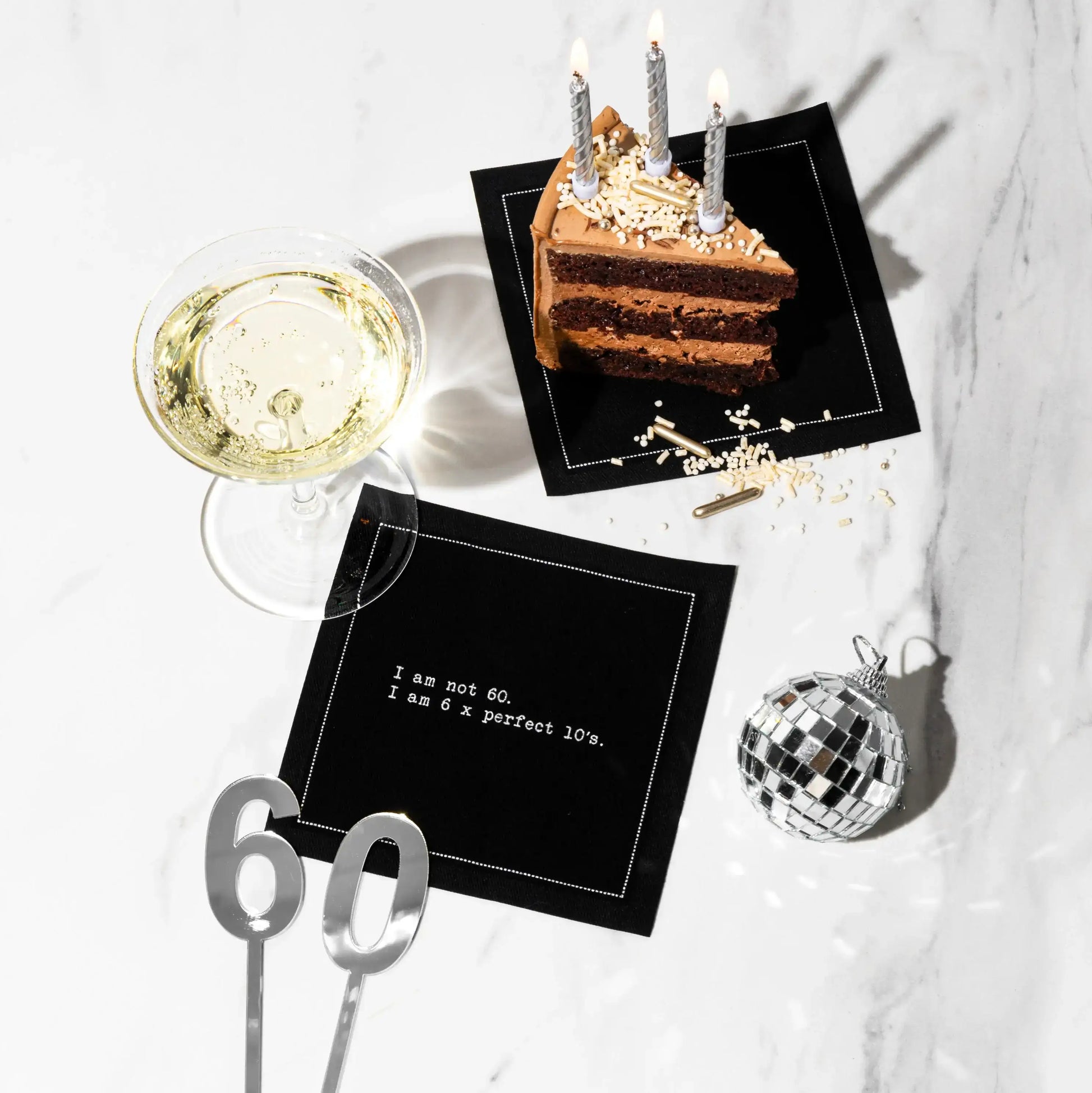 Organic cotton cocktail napkin, black, printed with the quote "I am not 60. I am 6 x perfect 10's," styled on a marble table with chocolate cake, champagne, and silver "60" cake toppers, from Five Star Napkins' 60th birthday quote collection
