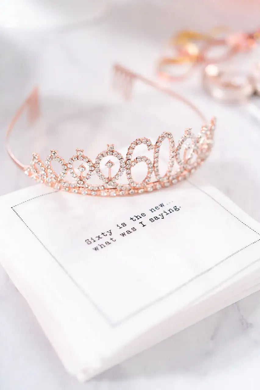 Organic cotton cocktail napkin printed with the quote "Sixty is the new... what was I saying," topped with a rose gold rhinestone "60" tiara, from Five Star Napkins' 60th birthday quote collection