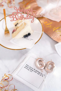 Organic cotton cocktail napkin printed with the quote "When you're 60, getting lucky means finding your car in the parking lot," styled with birthday cake, rose gold "60" candles, and party streamers, from Five Star Napkins' 60th birthday quote collection