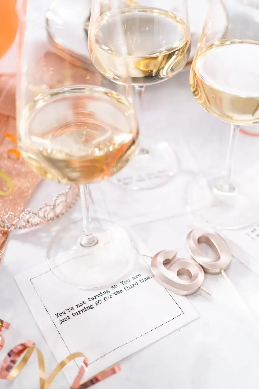 Organic cotton cocktail napkin printed with the quote "You're not turning 60. You are just turning 20 for the third time," styled on a celebration table with wine glasses and rose gold "60" candles, from Five Star Napkins' 60th birthday quote collection