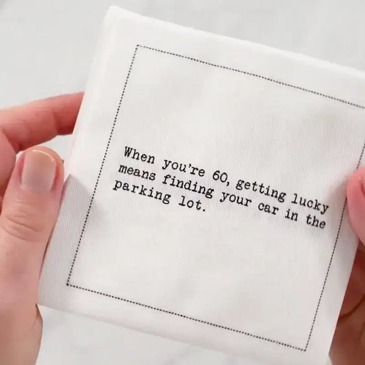 Hand holding an organic cotton cocktail napkin with black dotted border printed with the quote "When you're 60, getting lucky means finding your car in the parking lot," from Five Star Napkins' 60th birthday quote collection