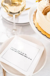 Organic cotton napkin with printed quote 'I've still got it at 70. I just can't remember where it is.' on a plate, next to a glass of white wine and a cake in a table setting.