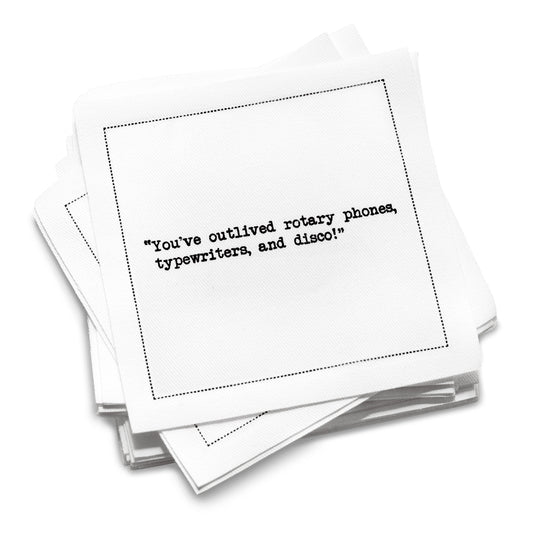 Organic cotton cocktail napkin with black dotted border printed with the quote "You've outlived rotary phones, typewriters, and disco!" from Five Star Napkins' 75th birthday quote collection