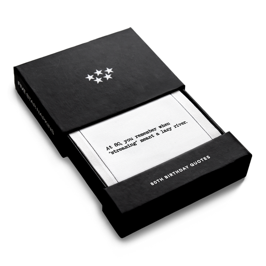 Open black gift box from Five Star Napkins' 80th birthday quote collection, revealing an organic cotton cocktail napkin printed with the quote "At 80, you remember when 'streaming' meant a lazy river,'"