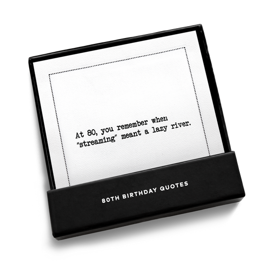 Black gift box from Five Star Napkins' 80th birthday quote collection, open to display an organic cotton cocktail napkin printed with the quote "At 80, you remember when 'streaming' meant a lazy river,'"