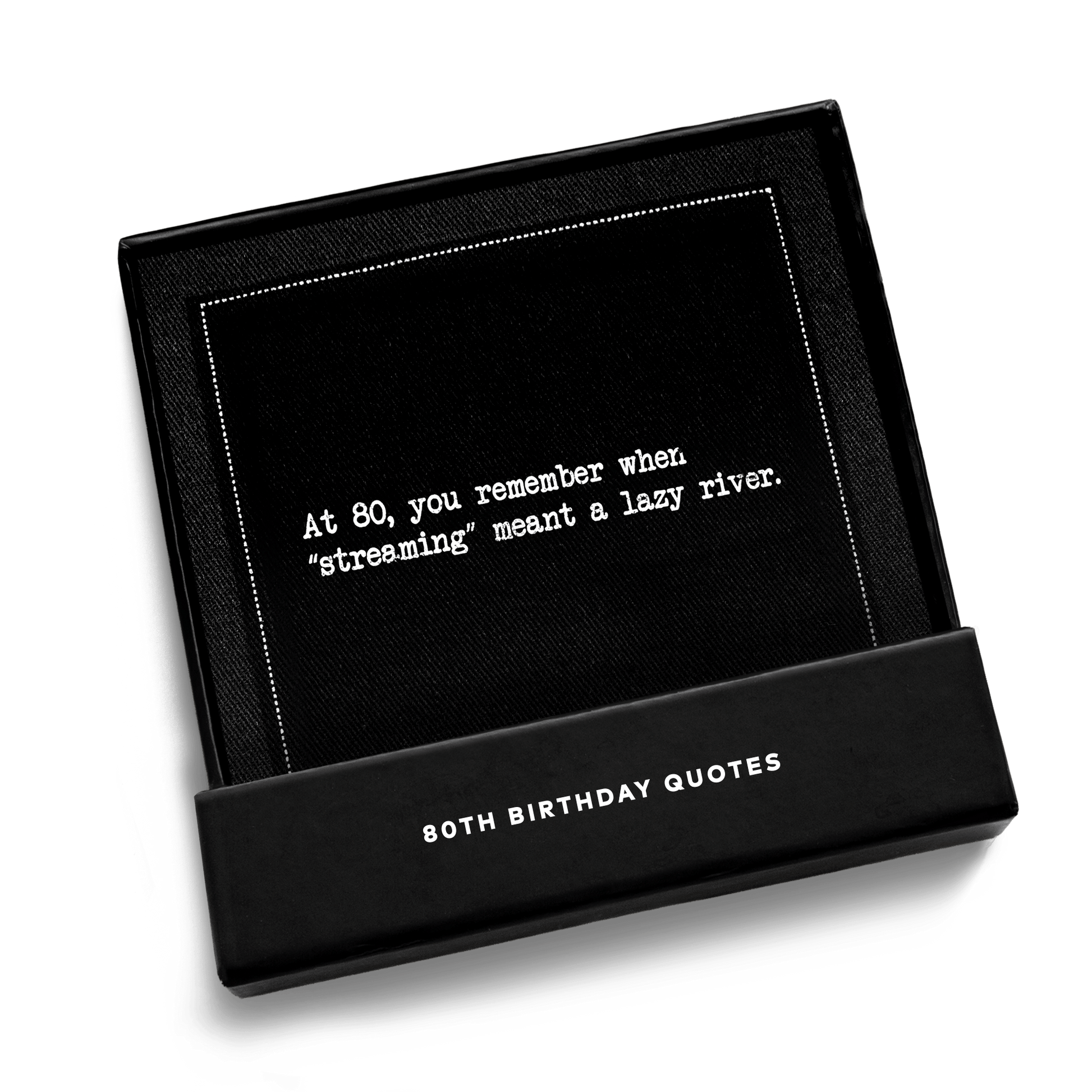 Open black gift box from Five Star Napkins' 80th birthday quote collection, revealing an organic cotton cocktail napkin, black, printed with the quote "At 80, you remember when 'streaming' meant a lazy river,'"