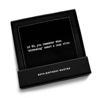 Open black gift box from Five Star Napkins' 80th birthday quote collection, revealing an organic cotton cocktail napkin, black, printed with the quote "At 80, you remember when 'streaming' meant a lazy river,'"