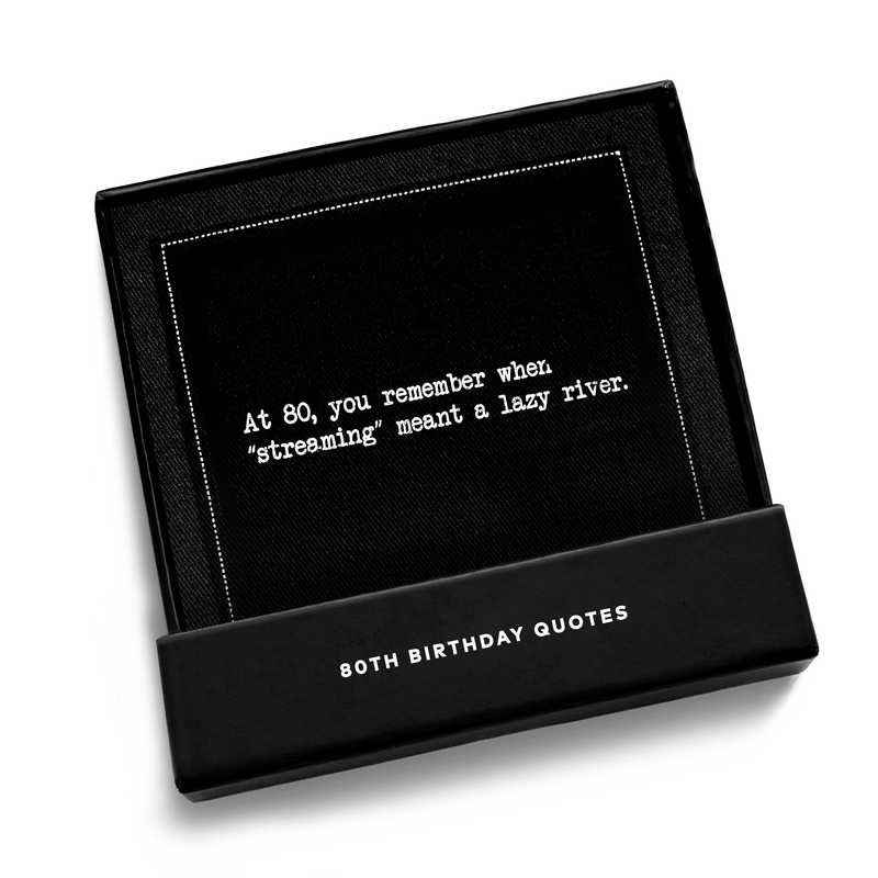 Open black gift box from Five Star Napkins' 80th birthday quote collection, revealing an organic cotton cocktail napkin, black, printed with the quote "At 80, you remember when 'streaming' meant a lazy river,'"