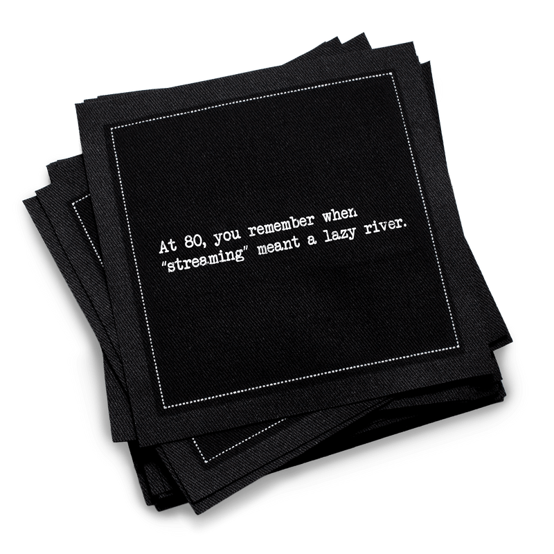 Organic cotton cocktail napkin, black, with white dotted border printed with the quote "At 80, you remember when 'streaming' meant a lazy river,'" from Five Star Napkins' 80th birthday quote collection